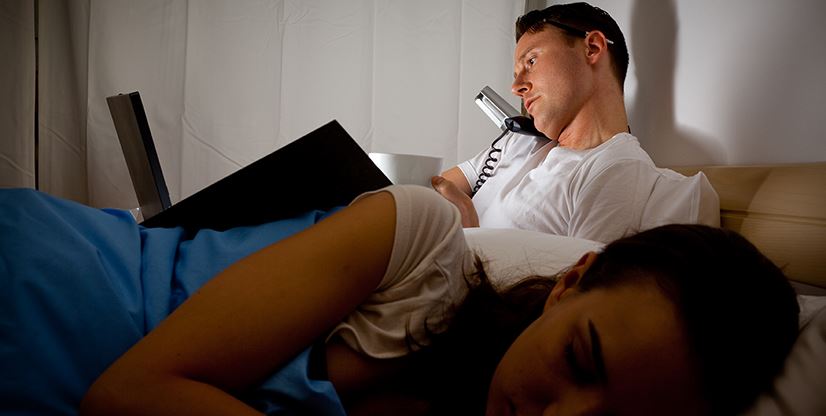 man on his laptop and on the phone in bed next to sleeping woman